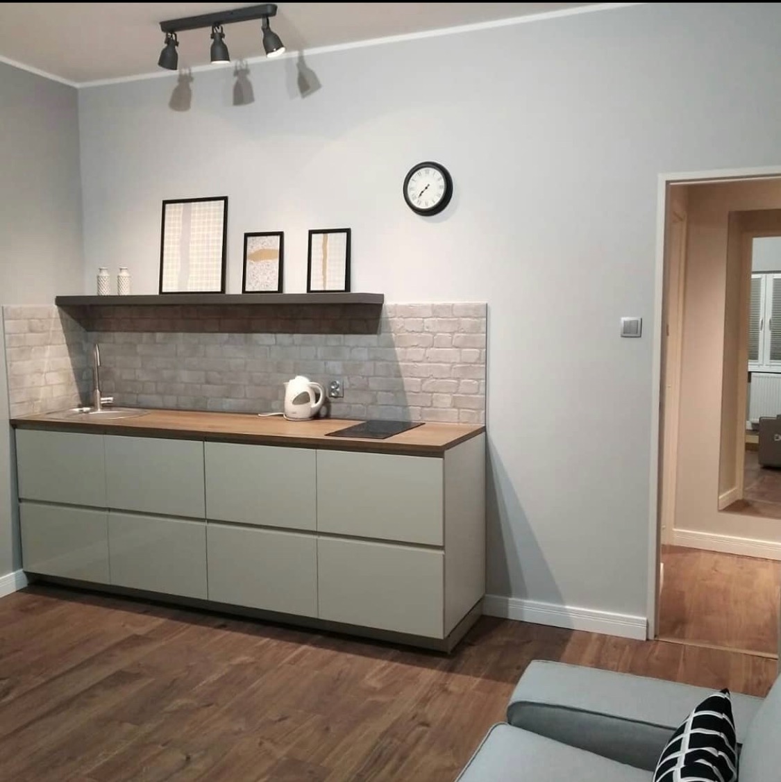 Nowoczesna kuchnia z szarymi szafkami, drewnianym blatem i ceglaną mozaiką na ścianie, uzupełniona czarnym zegarem i trzema obrazkami na półce, oświetlona reflektorami.