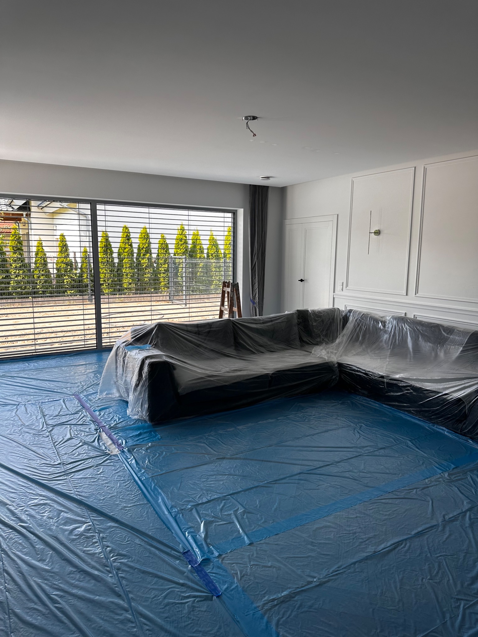 Pokój zabezpieczony folią malarską przed pracami wykończeniowymi, widoczna narożna sofa pod folią ochronną, niebieska folia na podłodze, okno z widokiem na zielone tuje, białe ściany...