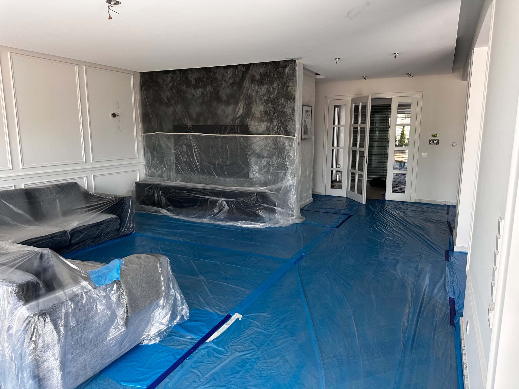 Salon zabezpieczony folią malarską i taśmą przed rozpoczęciem prac, meble zakryte folią ochronną, widoczne dekoracyjne listwy ścienne i przeszklone drzwi balkonowe.