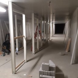 PHU ARSKOBUD - Długi korytarz w trakcie remontu, z ustawionymi pionowymi profilami konstrukcyjnymi, widoczne bloczki betonowe, drabina i narzędzia pracy.