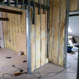 PHU ARSKOBUD - Ściana z metalowym stelażem wypełniona wełną mineralną, widoczne przewody elektryczne i narzędzie do cięcia na podłodze.