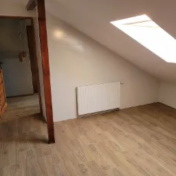 Poddasze z drewnopodobną podłogą, białymi ścianami wyłożonymi płytkami i oknem dachowym, widoczny drewniany słup i fragment otwartych drewnianych drzwi.