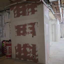 PHU ARSKOBUD - Ściana w trakcie szpachlowania z widocznymi łatami i niedoskonałościami, w tle rury wentylacyjne pod sufitem i materiały izolacyjne.