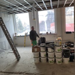 PHU ARSKOBUD - Wnętrze w trakcie prac wykończeniowych: widoczna metalowa konstrukcja sufitu podwieszanego, drabina oparta o ścianę, stosy wiader z farbą Caparol i Dekoral, oraz pracownik w zielonych spodniach...