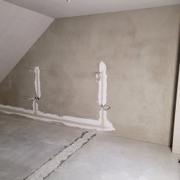 PHU ARSKOBUD - Surowe wnętrze poddasza w trakcie prac: ściany z częściowo nałożoną gładzią, bruzdy na kable elektryczne wypełnione gipsem, surowy beton na podłodze, widoczne przewody i puszki elektryczne.