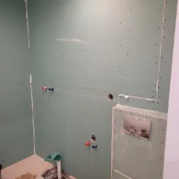 Wnętrze łazienki w trakcie prac: zielone płyty gipsowo-kartonowe na ścianach, widoczne instalacje wodne i kanalizacyjne, częściowo przygotowane podłoże pod posadzkę, stelaż podtynkowy WC...