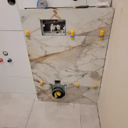 Montaż płytek imitujących marmur na ścianie z otworem rewizyjnym, z użyciem klipsów poziomujących, widoczne instalacje sanitarne w otworze, fragment ułożonej podłogi z płytek.