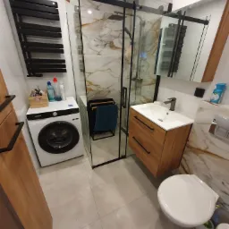 Nowoczesna łazienka z kabiną prysznicową typu walk-in z czarnymi profilami, umywalką z szafką z drewnopodobnymi frontami, białą pralką Samsung i grzejnikiem drabinkowym w kolorze czarnym.