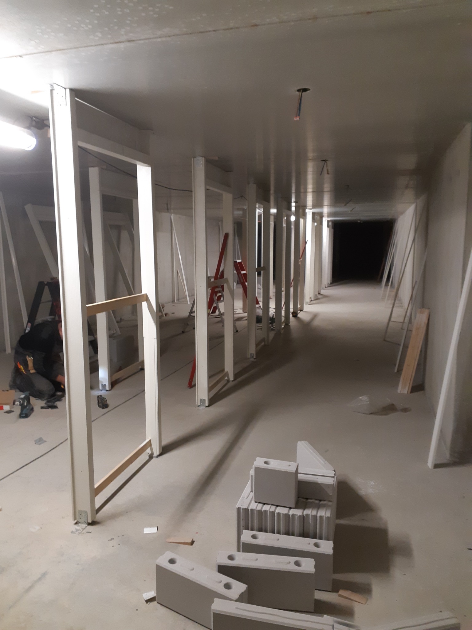 Długi korytarz w trakcie remontu, z ustawionymi pionowymi profilami konstrukcyjnymi, widoczne bloczki betonowe, drabina i narzędzia pracy.