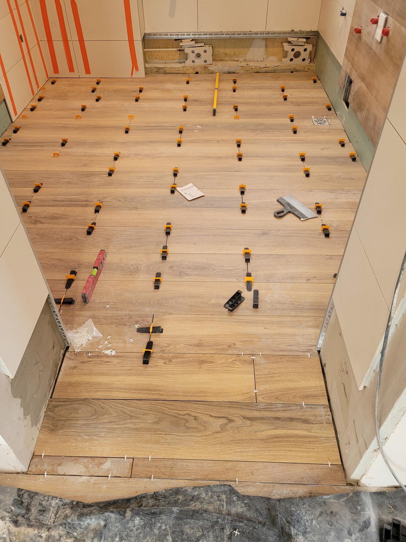 Podłoga w trakcie układania płytek imitujących drewno, z użyciem klipsów poziomujących i krzyżyków dystansowych, widoczna paca do rozprowadzania kleju i poziomica.