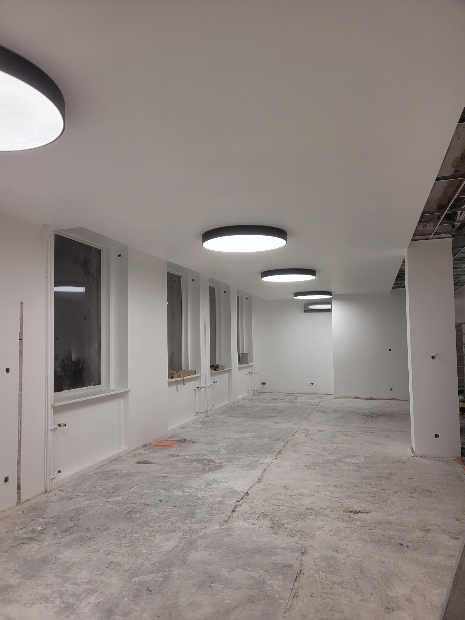 Wnętrze w trakcie prac przygotowawczych do wykończenia, widoczne surowe ściany, okna oraz betonowa podłoga, oświetlona okrągłymi lampami sufitowymi.