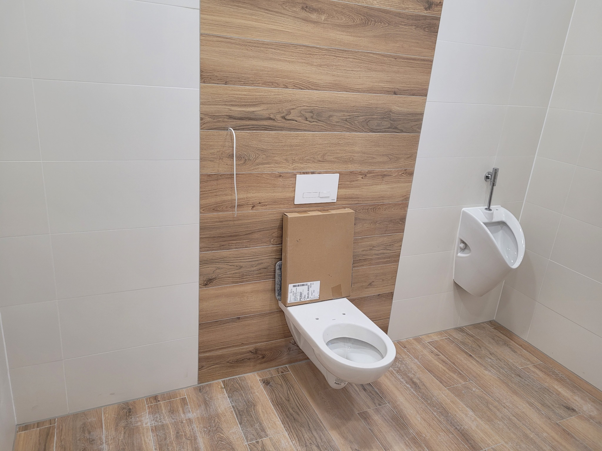 Wisząca miska WC w trakcie montażu, z kartonowym opakowaniem na górze, obok zamontowany pisuar, ściany wyłożone białymi i drewnopodobnymi płytkami, podłoga z paneli imitujących drewno.