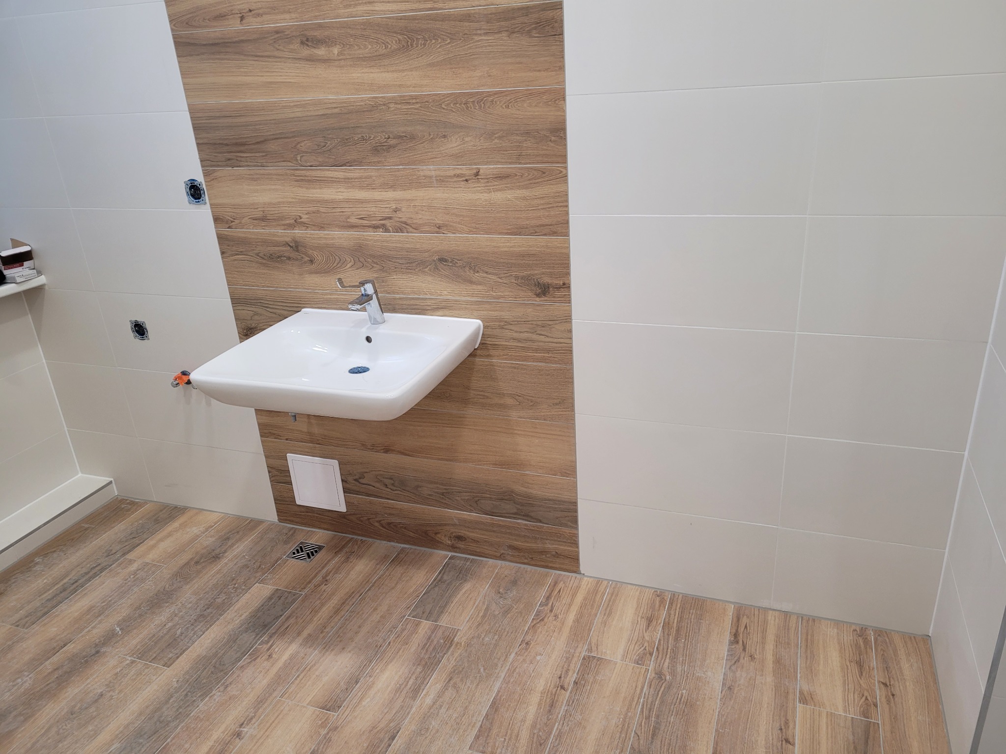 Nowoczesna łazienka z białymi płytkami i drewnopodobną okładziną ścienną oraz podłogą, wisząca biała umywalka z chromowaną baterią, kratka ściekowa w podłodze.