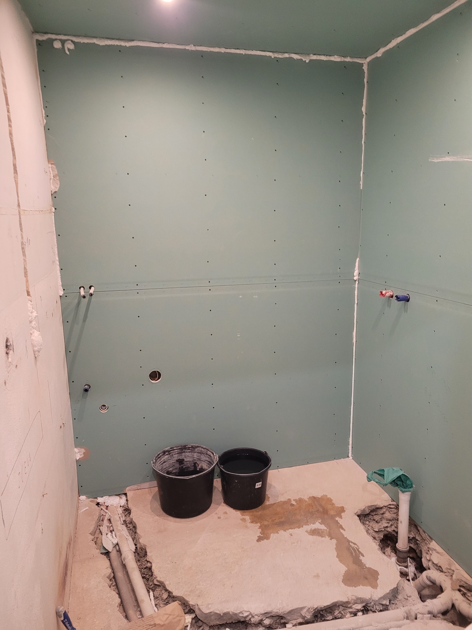 Łazienka w trakcie prac przygotowawczych: ściany wyłożone płytami gipsowo-kartonowymi, widoczne rury instalacji wodnej, dwa czarne wiadra na podłodze i odsłonięte fragmenty posadzki wokół rur...