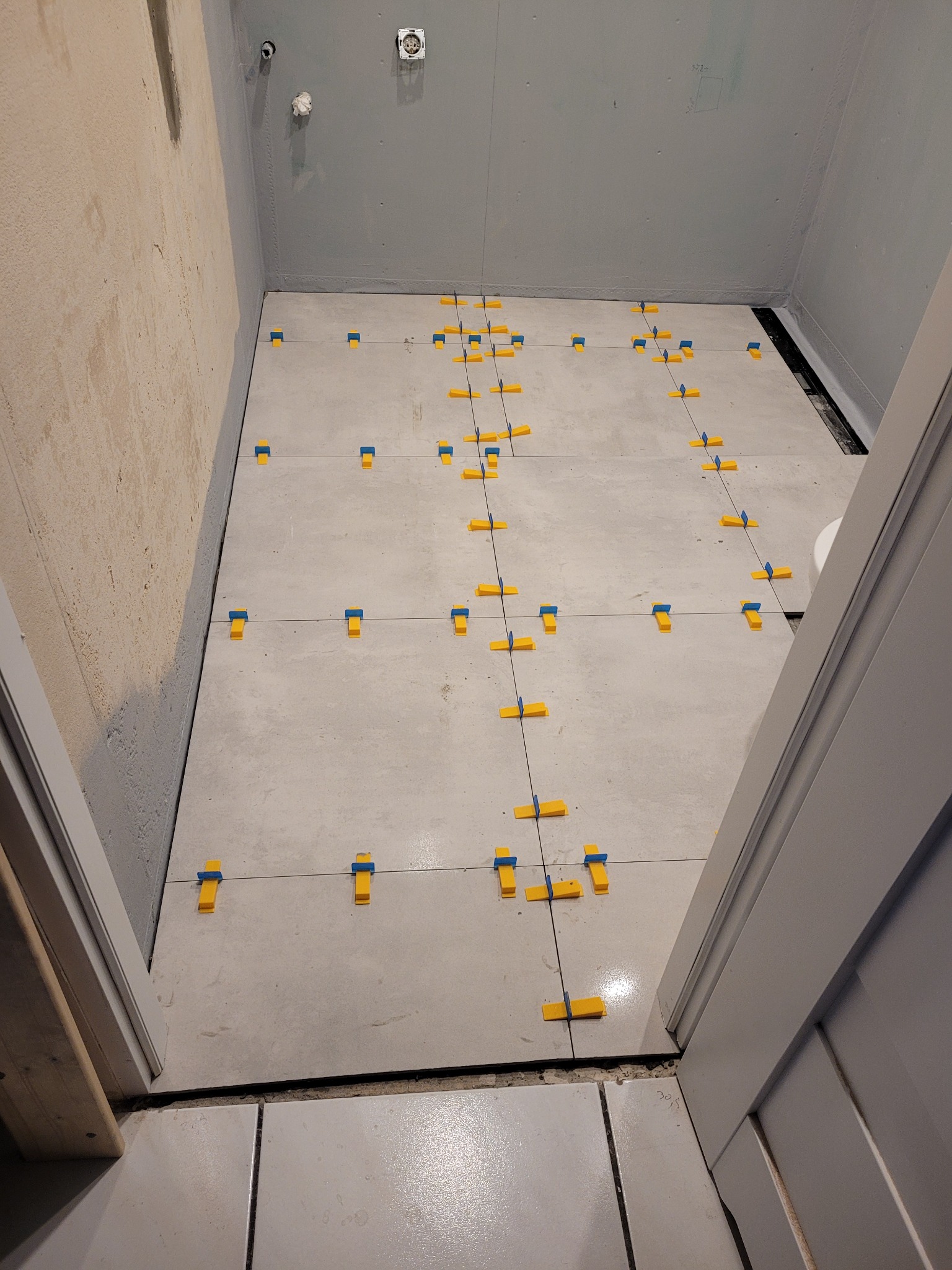 Układanie szarych płytek podłogowych w łazience, widoczne krzyżyki dystansowe w kolorze żółto-niebieskim, ściany przygotowane pod wykończenie, widok zza ościeżnicy.