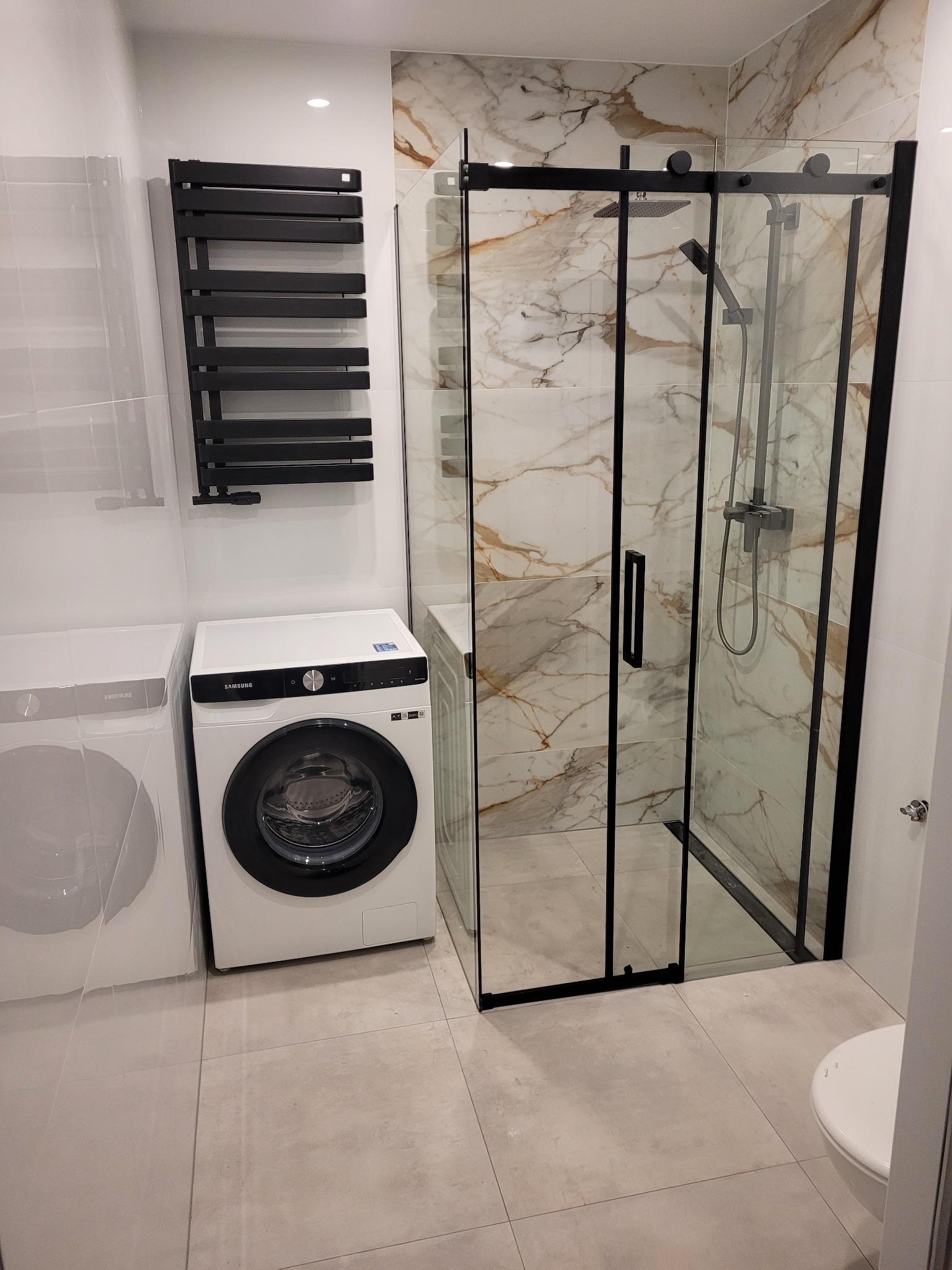 Nowoczesna łazienka z kabiną prysznicową z czarnymi ramami, grzejnikiem drabinkowym, marmurowymi płytkami i dwiema pralkami Samsung.