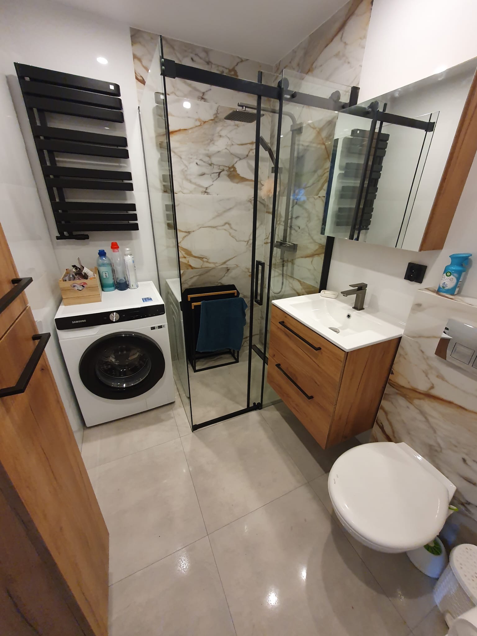 Nowoczesna łazienka z kabiną prysznicową typu walk-in z czarnymi profilami, umywalką z szafką z drewnopodobnymi frontami, białą pralką Samsung i grzejnikiem drabinkowym w kolorze czarnym.