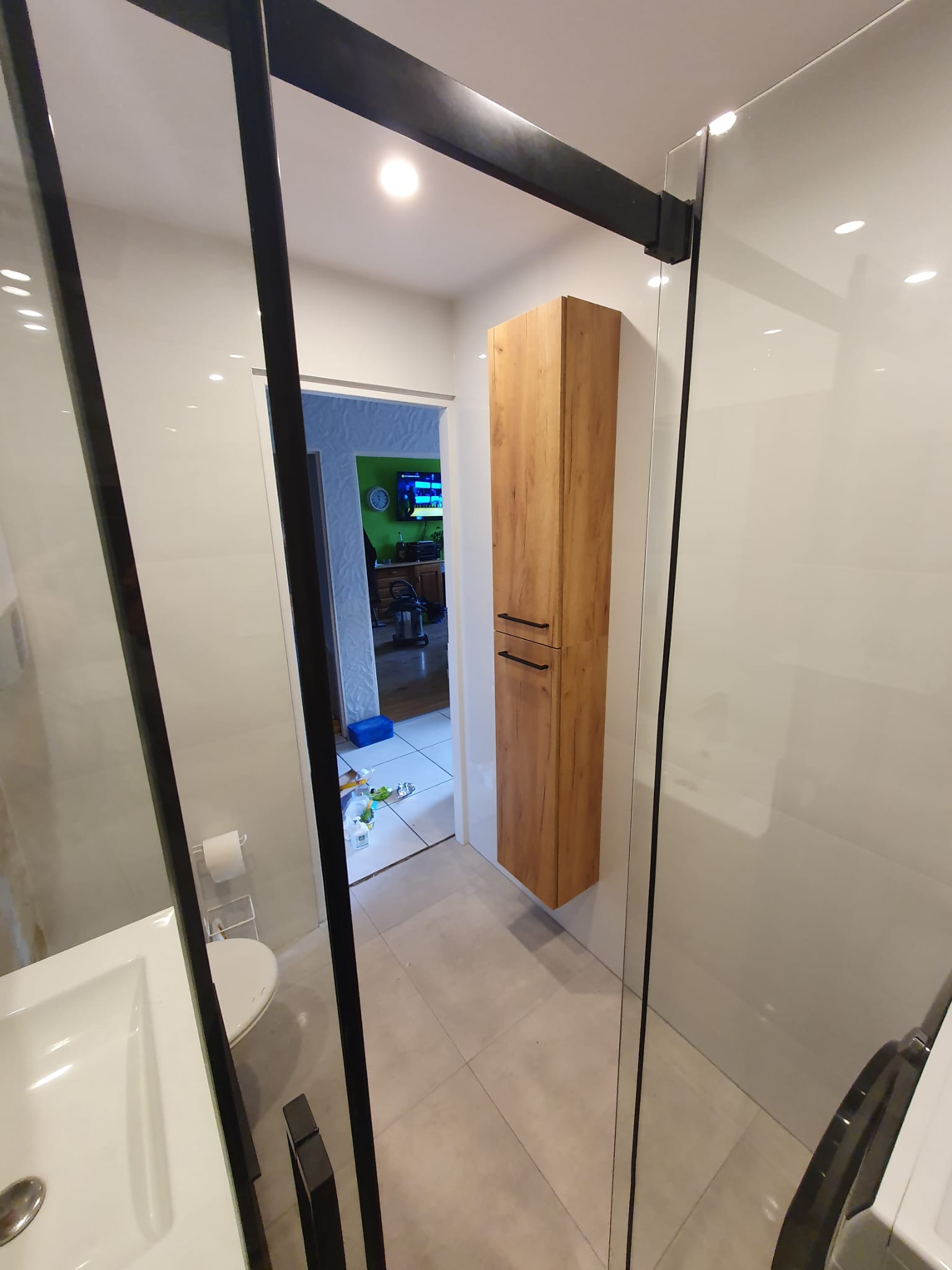 Nowoczesna łazienka z kabiną prysznicową typu walk-in z czarnymi profilami, wiszącą szafką z drewnopodobnym frontem i białymi płytkami wielkoformatowymi na ścianach.