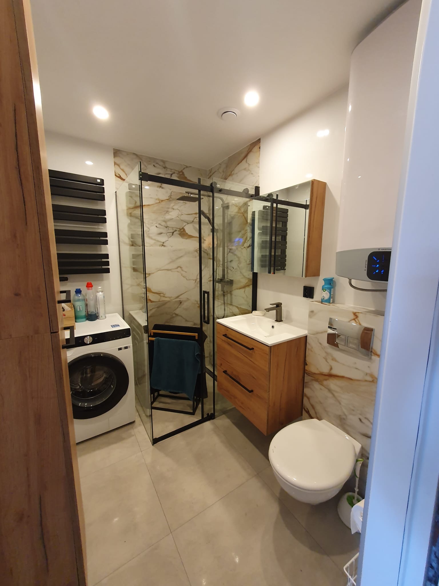 Nowoczesna łazienka z kabiną prysznicową typu walk-in z czarnymi ramami, drewnianą szafką pod umywalką, wiszącą toaletą i pralką w rogu.