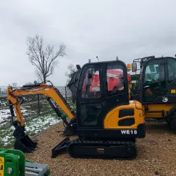 Żółto-czarna mini koparka WOLF WE18 na żwirowym podłożu, obok widoczna ładowarka teleskopowa Massey Ferguson i zielony element maszyny rolniczej, w tle drzewo bez liści i resztki śniegu.