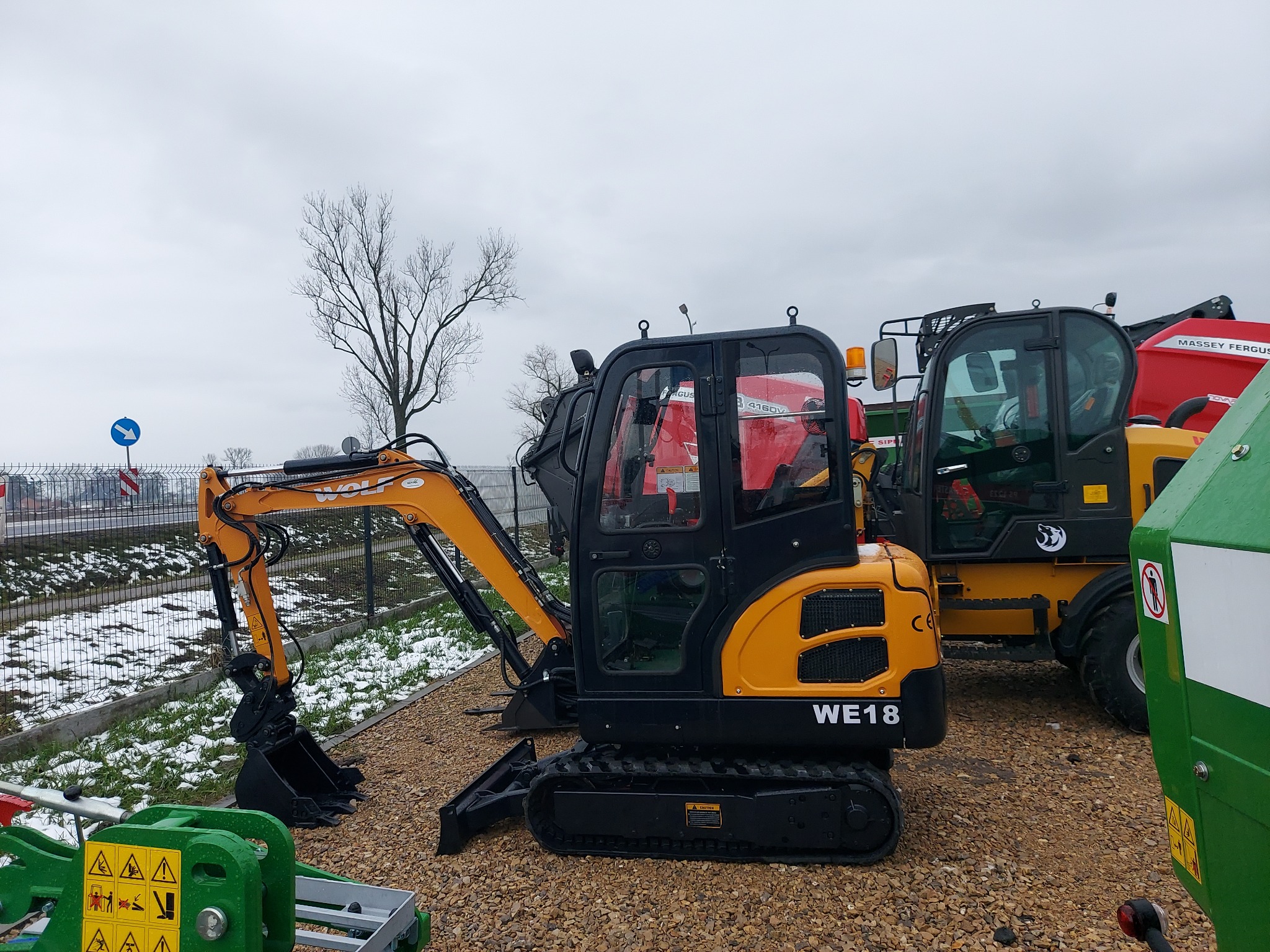 Żółto-czarna mini koparka WOLF WE18 na żwirowym podłożu, obok widoczna ładowarka teleskopowa Massey Ferguson i zielony element maszyny rolniczej, w tle drzewo bez liści i resztki śniegu.
