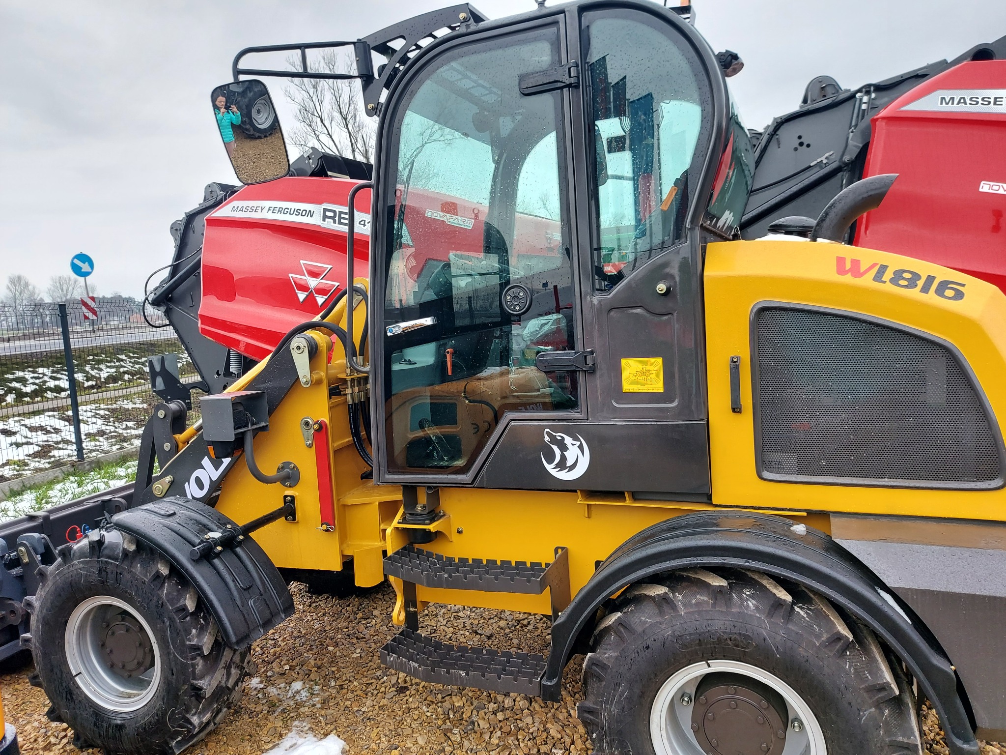 Żółto-czarna ładowarka teleskopowa W1816 z elementami czerwonymi marki Massey Ferguson, zaparkowana na żwirowym podłożu, z widocznymi oponami i kabiną z kroplami deszczu, na tle ogrodzenia...