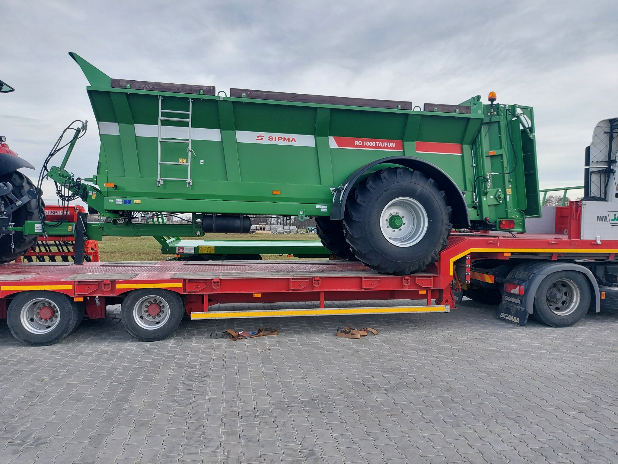 Zielona przyczepa rolnicza SIPMA RO 1000 Tajfun, przewożona na czerwonej platformie niskopodwoziowej ciągniętej przez ciągnik Scania, na szarym brukowanym placu.