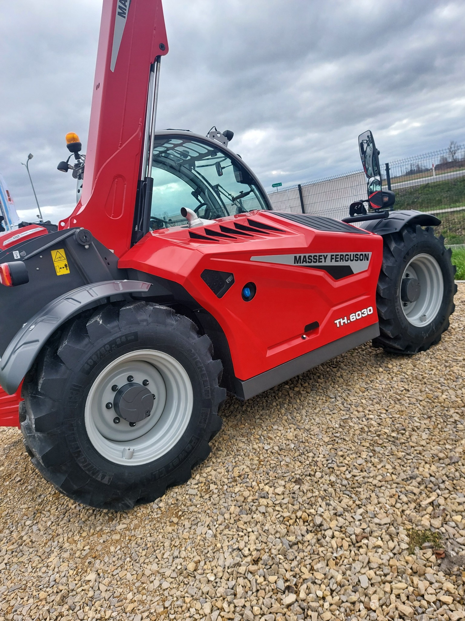 Czerwona ładowarka teleskopowa Massey Ferguson TH.6030 na żwirowym podłożu, widok z boku, pochmurne niebo w tle.