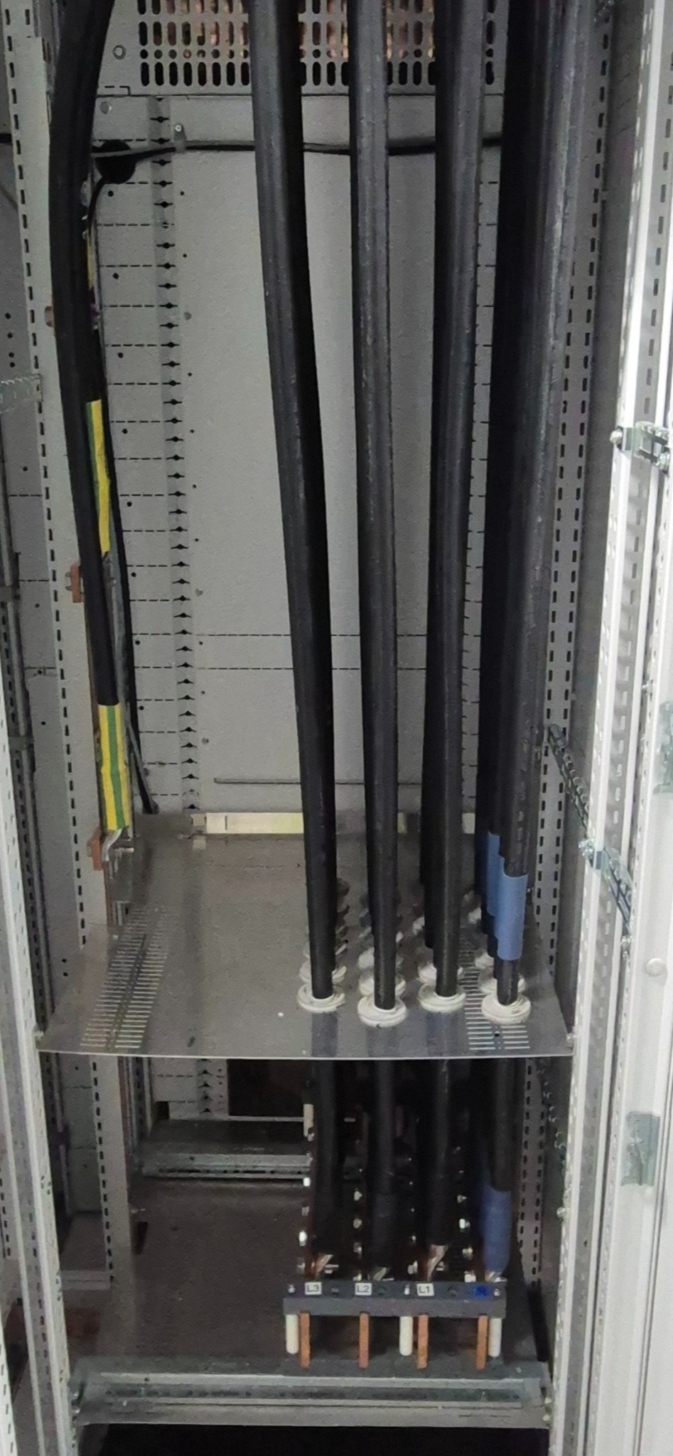 Kable elektryczne w szafie sterowniczej, widoczne połączenia i oznaczenia faz. Kable ułożone pionowo, prowadzone przez metalową płytę wewnątrz szafy.