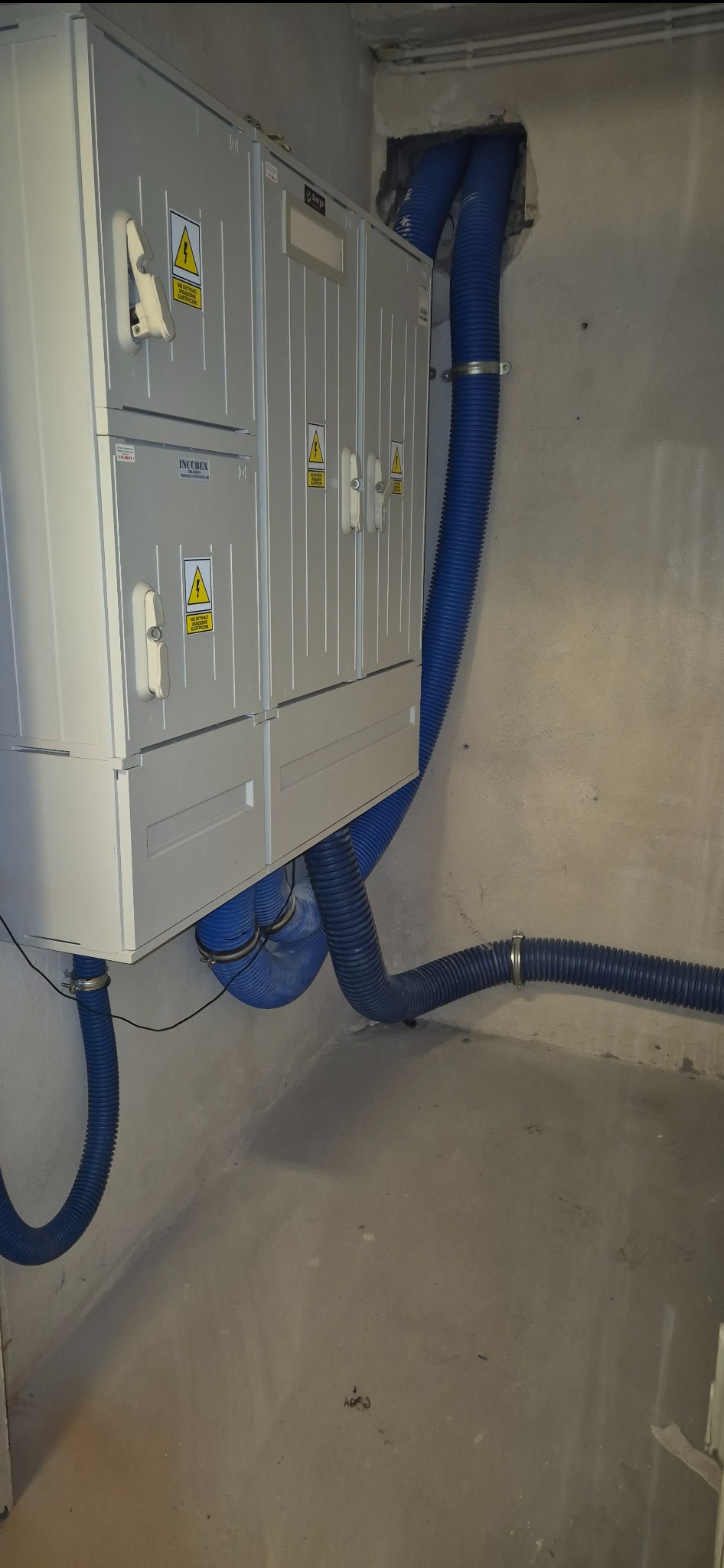 Rozdzielnica elektryczna IncoBex z niebieskimi rurami karbowanymi w surowym wnętrzu. Widoczne oznaczenia ostrzegawcze i elementy montażowe.