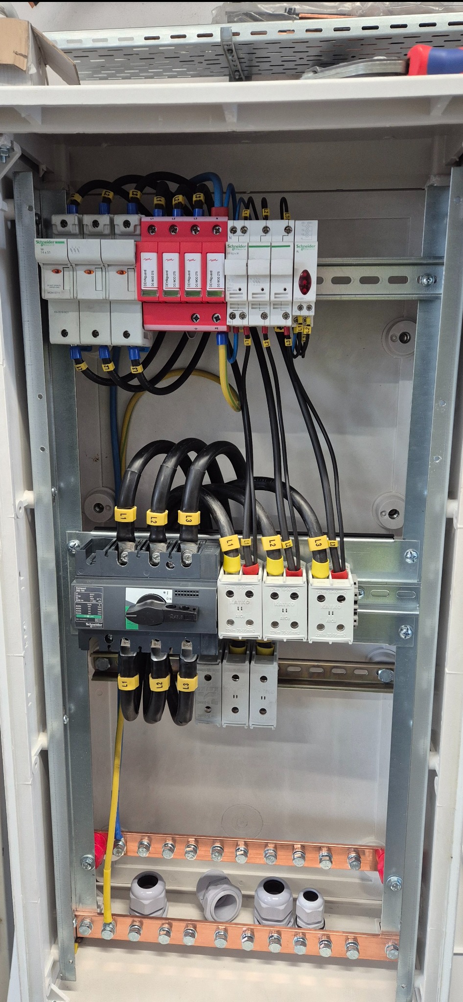 Wnętrze skrzynki elektrycznej z zainstalowanymi bezpiecznikami, przewodami oznaczonymi żółtymi etykietami i zabezpieczeniami przeciwprzepięciowymi w kolorze czerwonym.