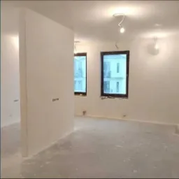 Pokój w trakcie prac wykończeniowych: ściany i sufit po nałożeniu gładzi, widoczne puszki elektryczne, surowy beton na podłodze, dwa okna z widokiem na budynek.