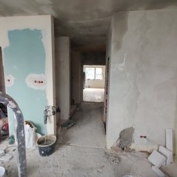 Michał Radzikowski - Wnętrze w trakcie prac wykończeniowych: ściany częściowo pokryte gładzią, widoczne instalacje elektryczne, mieszadło do zaprawy, białe bloczki i narzędzia budowlane na podłodze, perspektywa...