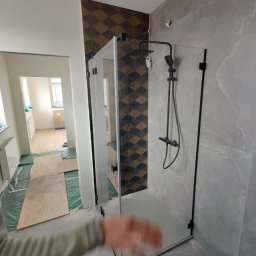 Michał Radzikowski - Nowoczesna kabina prysznicowa z czarną armaturą i szklanymi drzwiami na tle płytek w odcieniach szarości i wzorzystej ściany w trakcie remontu łazienki.