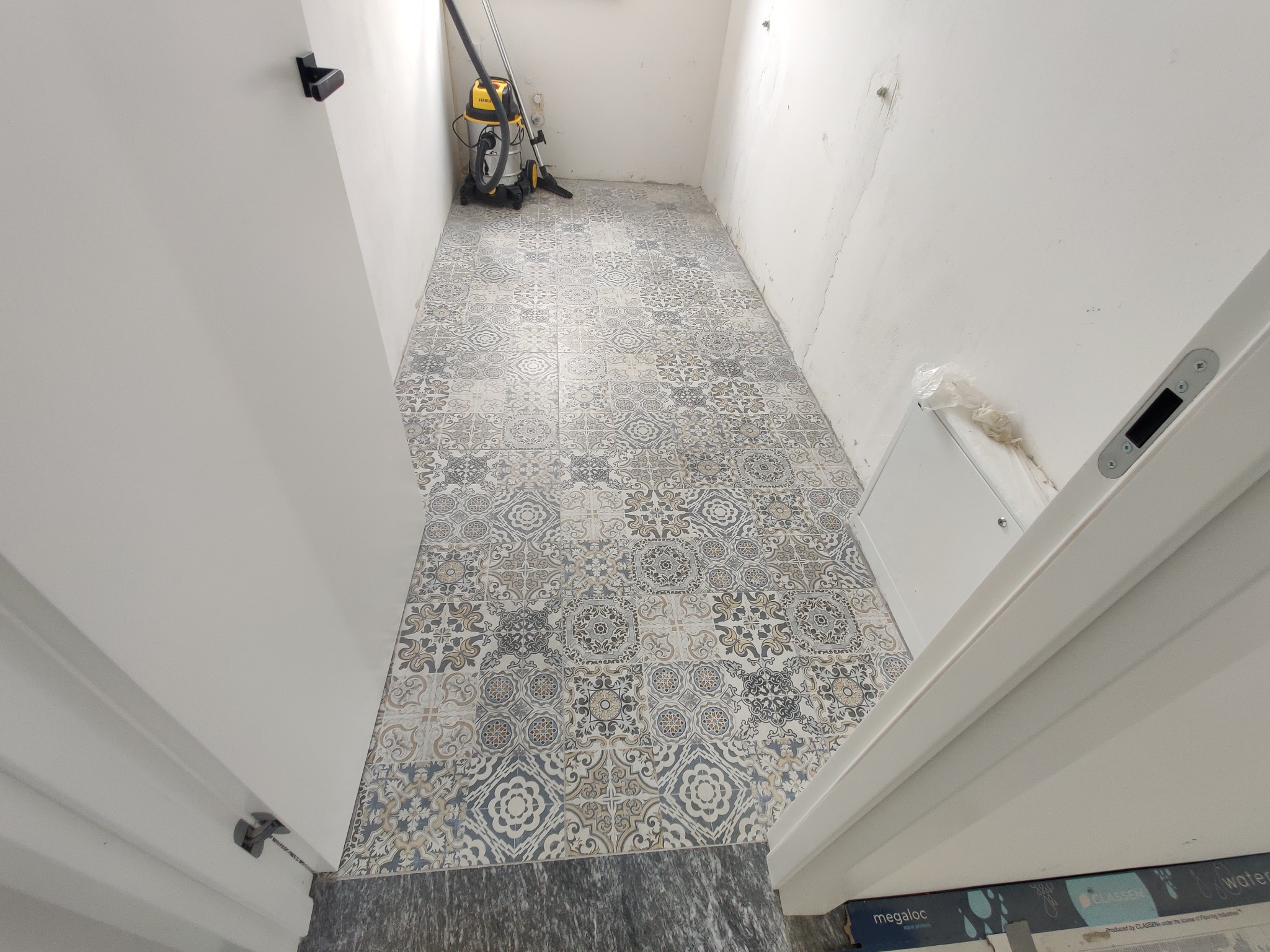 Nowo położona podłoga z płytek ceramicznych o wzorze patchworkowym w wąskim pomieszczeniu, widoczny odkurzacz przemysłowy i niedoskonałości na ścianach.