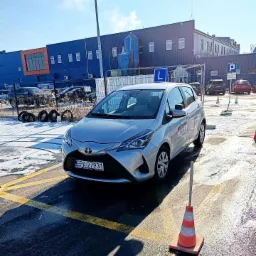 Srebrny Toyota Yaris z literą 'L' na dachu, zaparkowany na placu manewrowym z widocznymi liniami i pachołkami, na tle niebieskiego budynku przemysłowego.