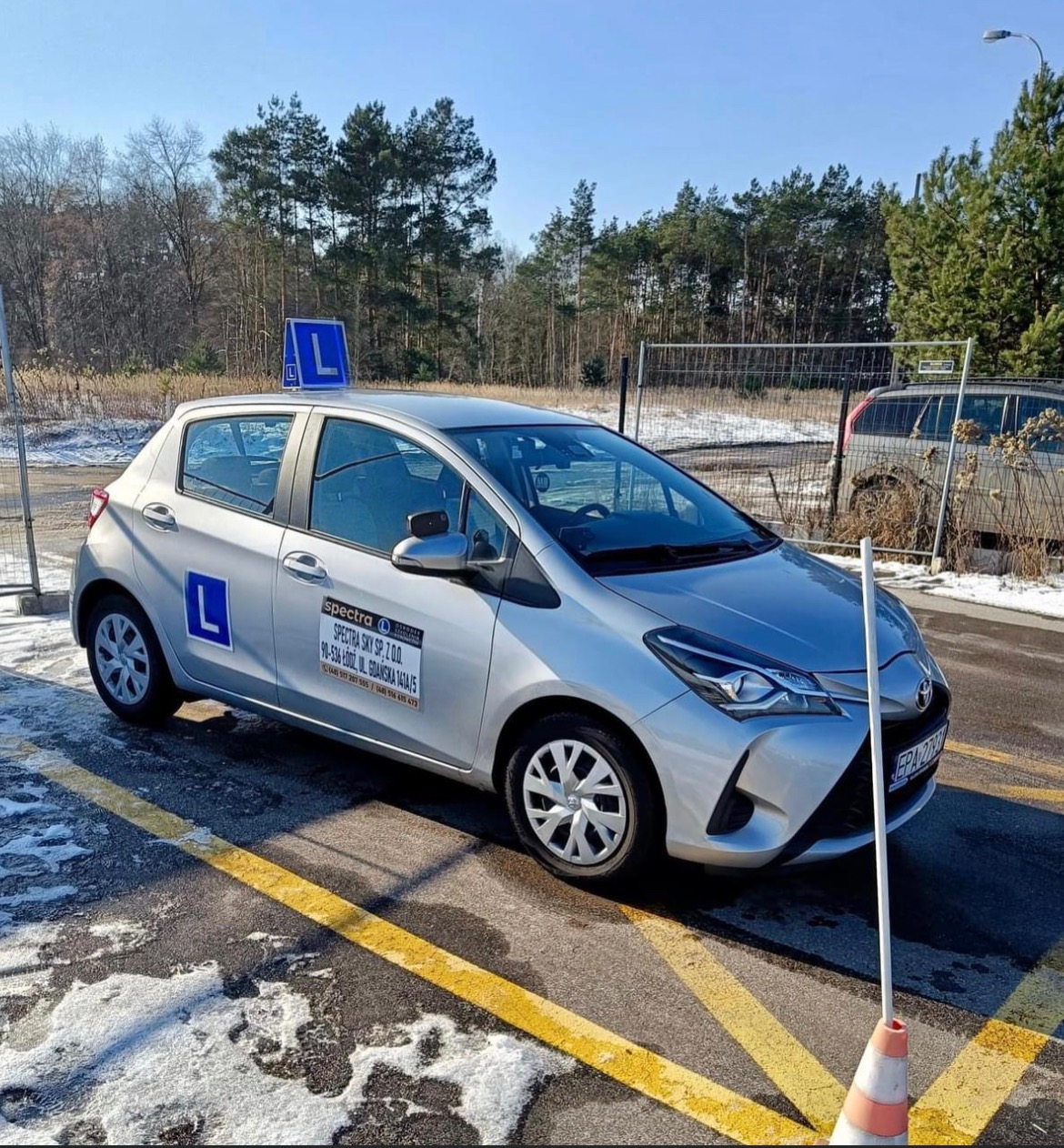 Srebrny samochód z naklejką i literą 'L' na dachu, zaparkowany na placu manewrowym z widocznym pachołkiem i resztkami śniegu.