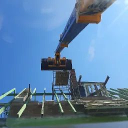 Demontaż dachu budynku z użyciem dźwigu, widok z dołu na rozbiórkę konstrukcji, fragmenty cegieł i drewnianych belek na tle błękitnego nieba.