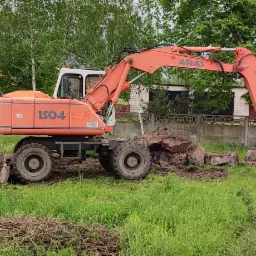 Pomarańczowa koparka Atlas 1504 na trawiastym terenie z pryzmą ziemi i stertą kamieni, w tle zabudowa jednorodzinna i drzewa.
