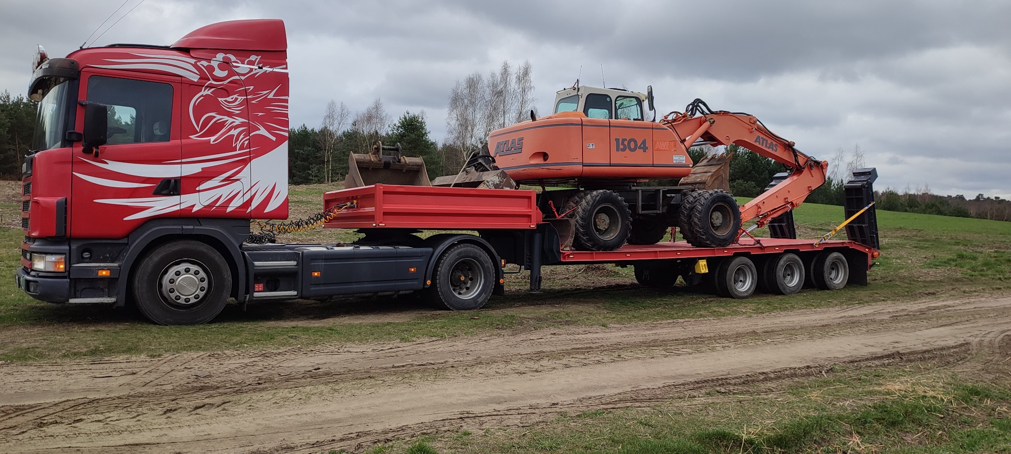 Czerwony ciągnik siodłowy Scania z grafiką orła transportuje na niskopodwoziowej platformie pomarańczową koparkę kołową Atlas 1504 na tle zielonej łąki i pochmurnego nieba.