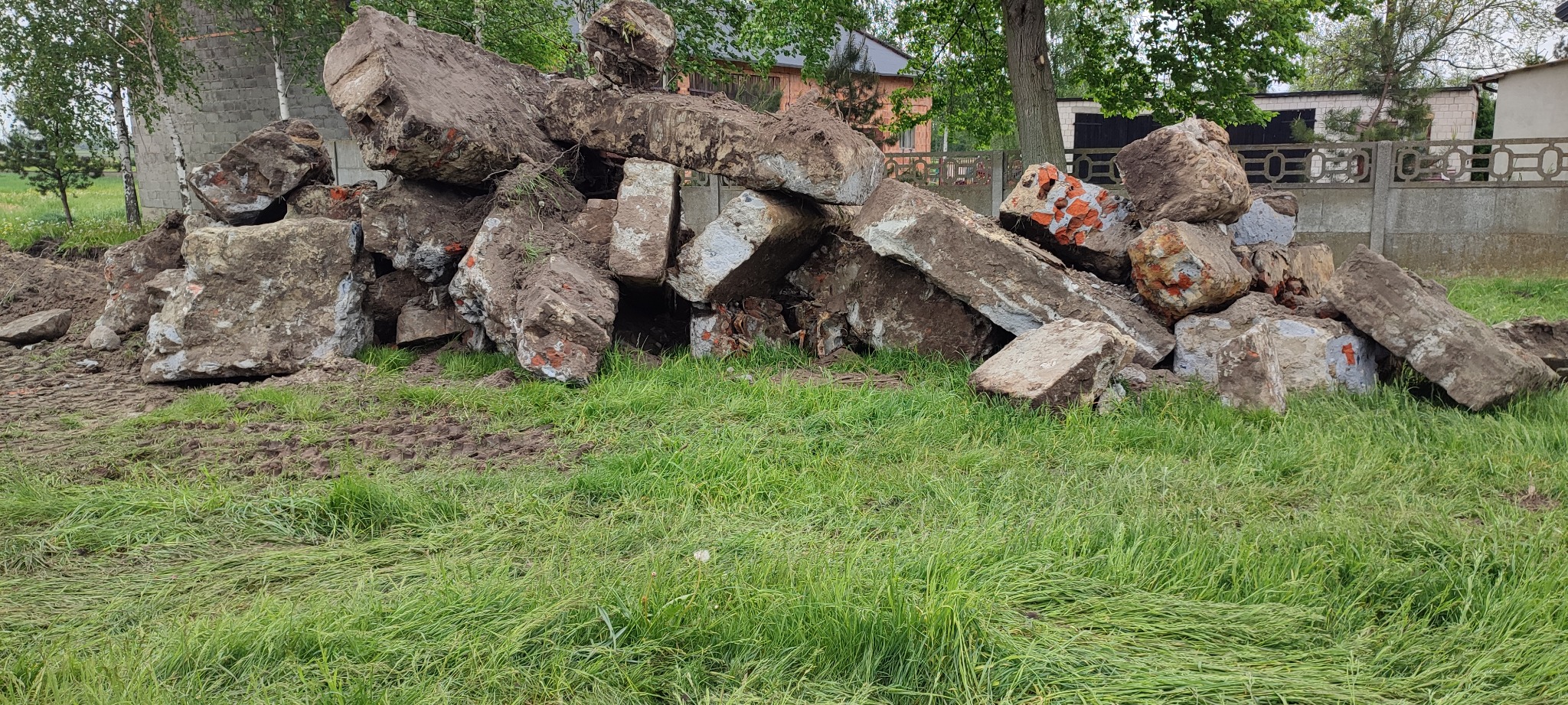 Stos betonowych bloków z resztkami cegieł i ziemi, leżący na trawie przed betonowym ogrodzeniem, po rozbiórce fundamentów.