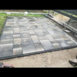 Ułożona, prostokątna nawierzchnia z szarej kostki brukowej z czarną obwódką, widok z góry, obok ziemia i trawa.