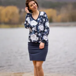 Kobieta w kwiecistej bluzce i granatowej sp&oacute;dnicy pozuje na piaszczystej plaży nad jeziorem, z zalesionymi wzg&oacute;rzami w tle.