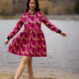 Kobieta w kwiecistej sukience z kapturem i niebieskich trampkach, stojąca na piaszczystej plaży z jeziorem i zalesionymi wzgórzami w tle.