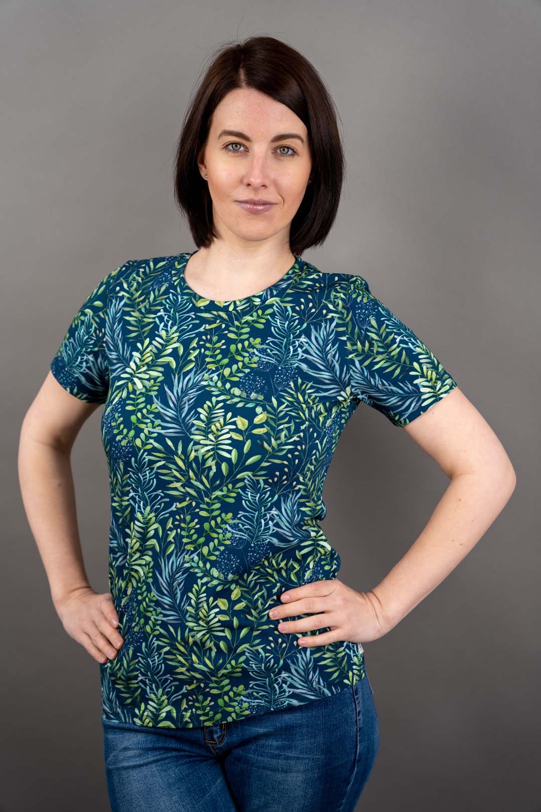 Kobieta w bluzce z motywem liści na tle szarego studia, ręce na biodrach, prezentacja produktu odzieżowego.
