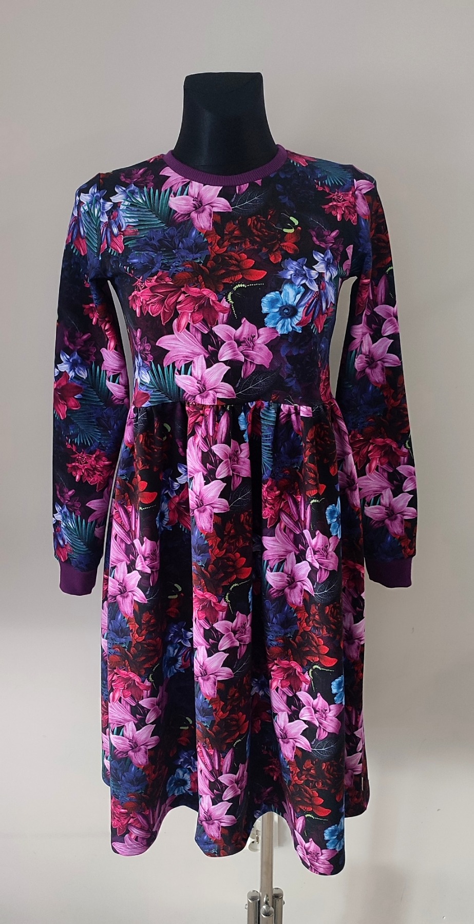 Sukienka z długim rękawem, uszyta z czarnego materiału w bogaty, tropikalny wzór kwiatowy w odcieniach różu, czerwieni i niebieskiego, prezentowana na manekinie.