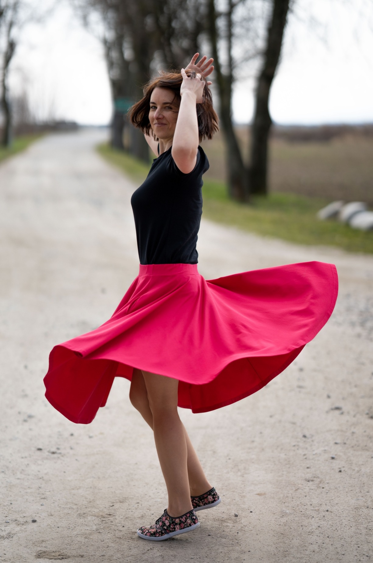 Kobieta w czarnej koszulce i rozkloszowanej, jaskrawoczerwonej spódnicy wiruje na polnej drodze, unosząc ręce do góry.