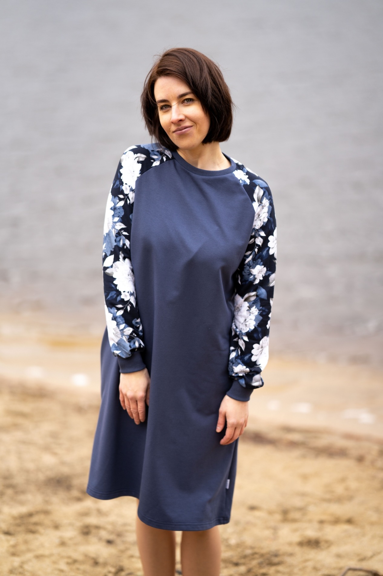 Kobieta w granatowej sukience z rękawami w kwiaty na tle plaży i wody.