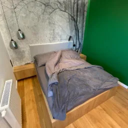 Drewniane łóżko z szarą pościelą i zagłówkiem, drewniana podłoga, ściana z fototapetą z motywem drzew i zielona ściana, lampki nocne wiszące.