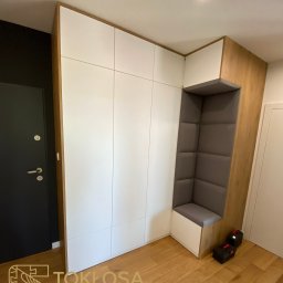 STOKŁOSA - Meble na wymiar RAFAŁ STOKŁOSA - Biała szafa wnękowa z drewnianymi akcentami i siedziskiem z szarej tapicerki, wkomponowana w przedpokój. Widoczne narzędzia na podłodze.